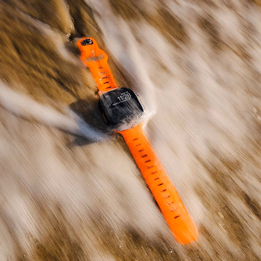 Apple Watch з помаранчевим ремінцем Nomad Sport Band Ultra Orange (41/42 мм) у морській піні та на піску.