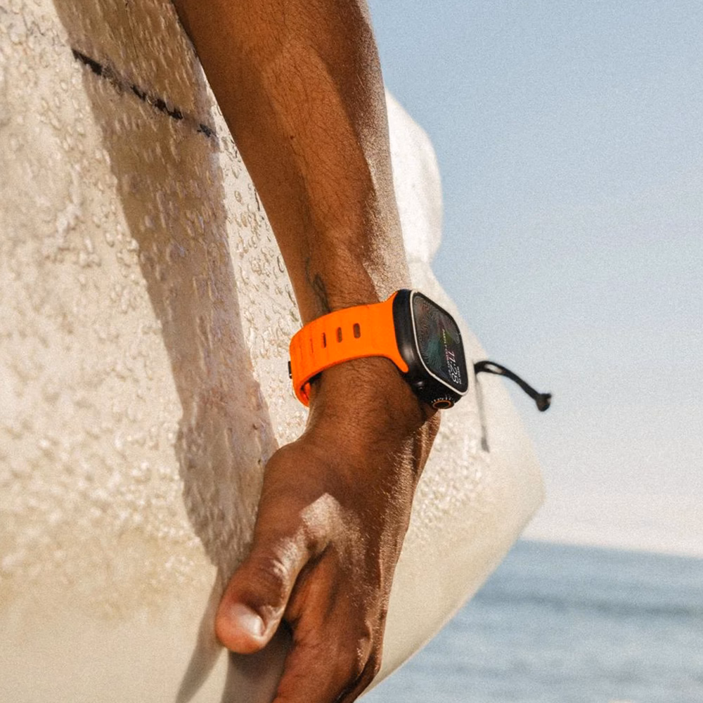 Чоловіча рука з Apple Watch у помаранчевому ремінці Nomad Sport Band Ultra Orange тримає серфборд на пляжі.